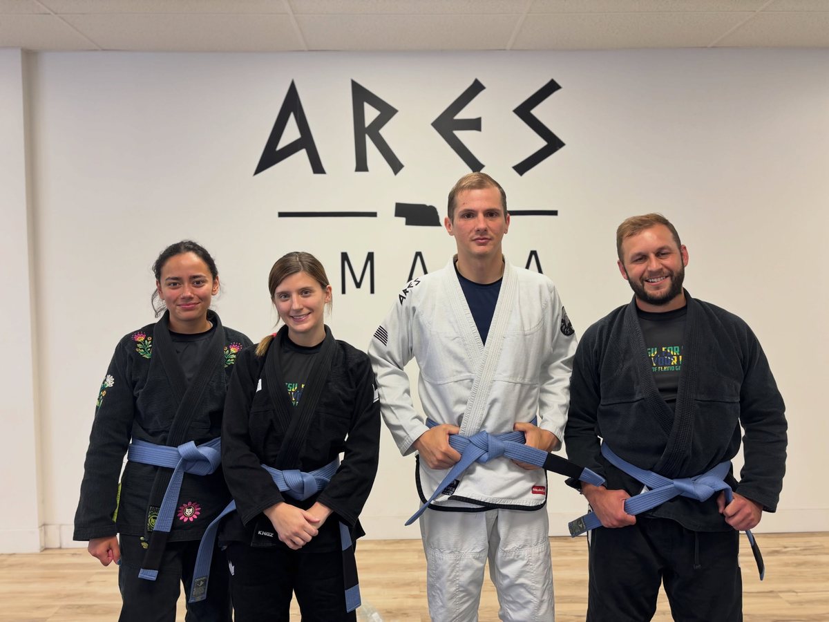 Four Ares BJJ Omaha students in gis with their new blue belts at the Ares wall
